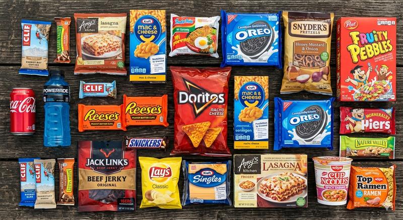 Flat lay of common processed foods with hidden added sugar including chips, cereal, candy bars, frozen meals, and packaged snacks on a dark wooden surface
